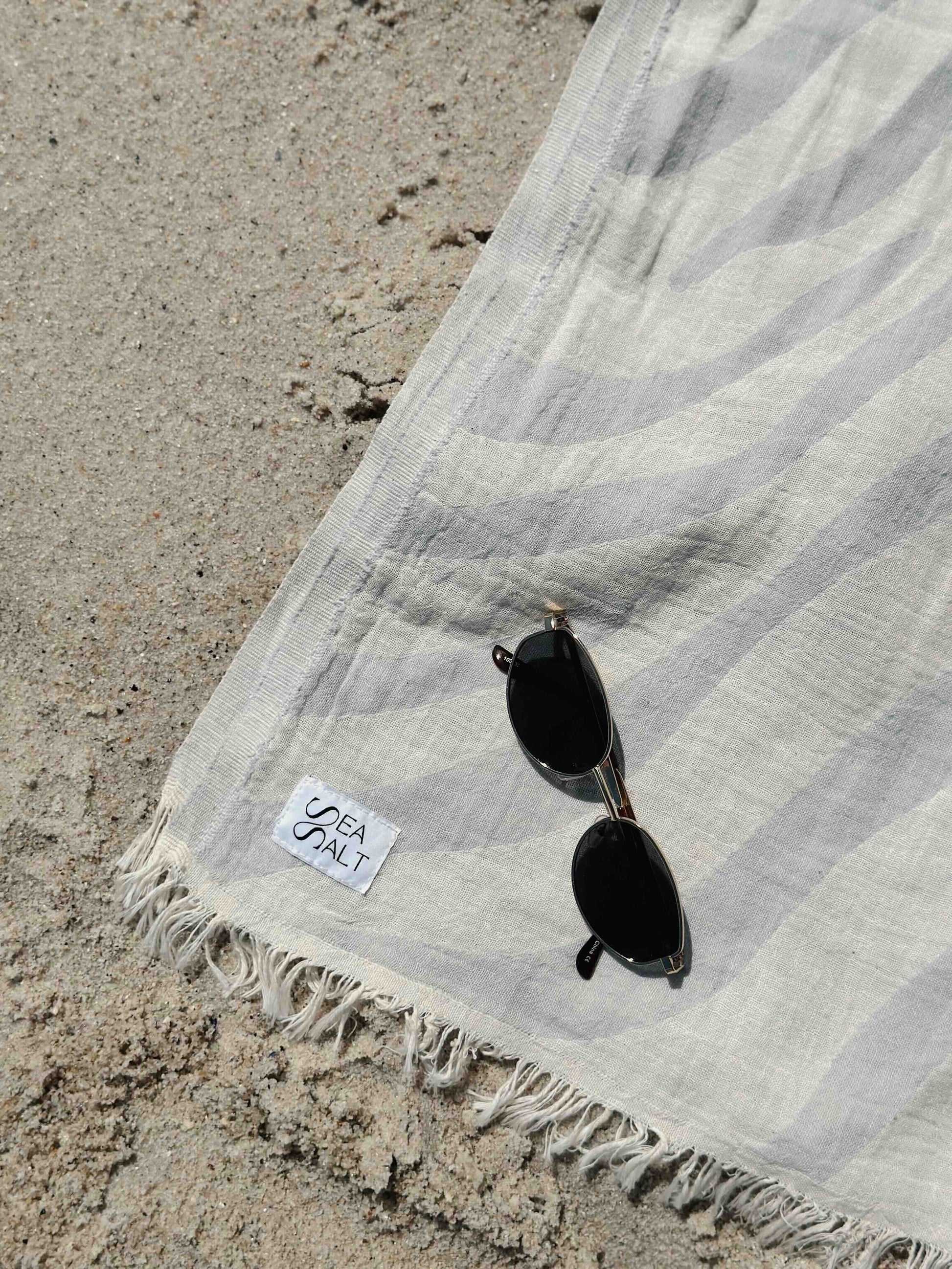 unfolded light blue beach towel in the sand with sea salt logo and sun glasses