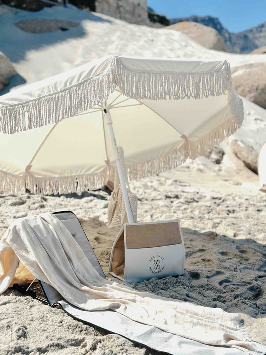 beach chair and umbrella with a cooler bag and beach towel on the beach with white sand